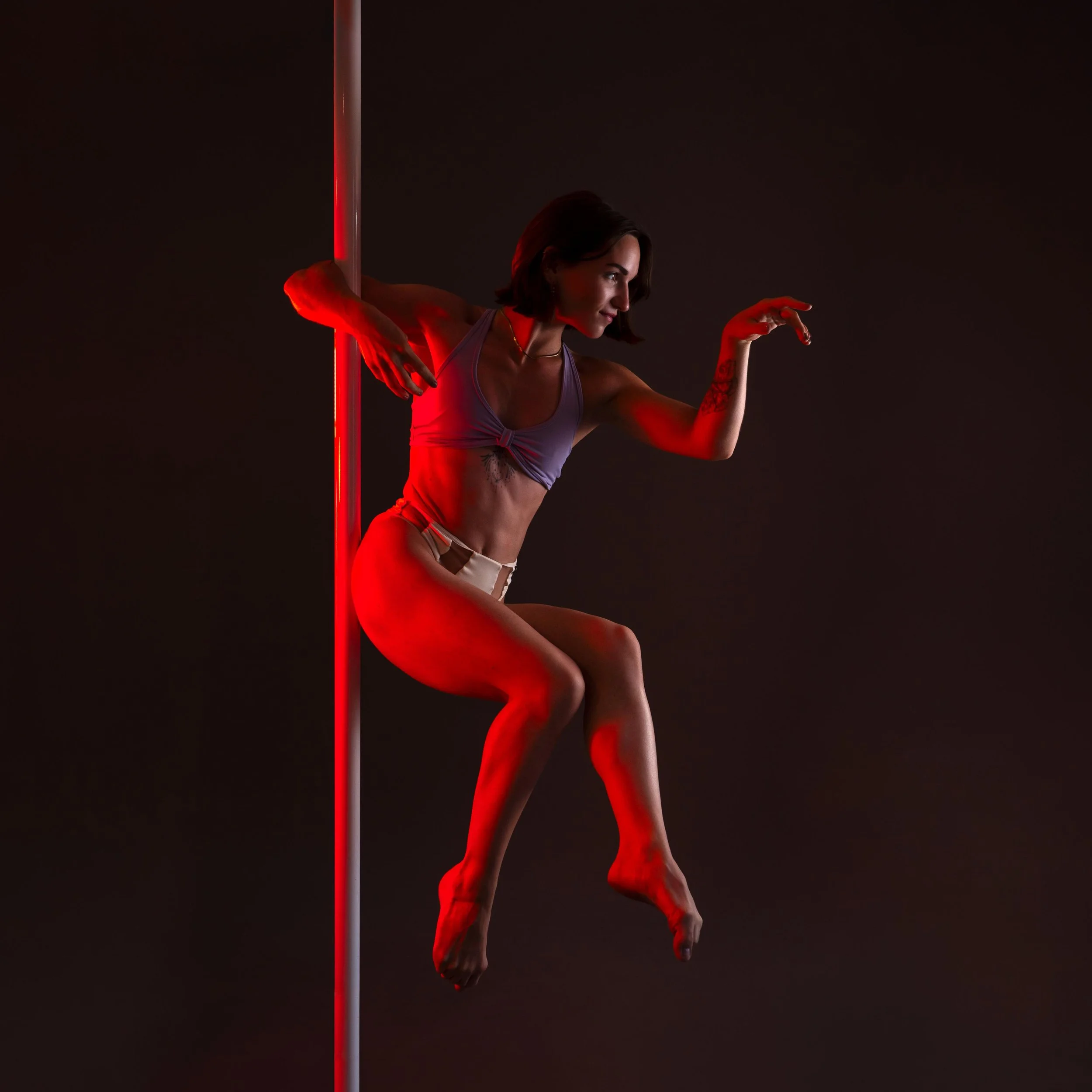 Une femme effectuant une pose de danse ou d'exercice dans un environnement sombre avec un éclairage rouge. Elle est accrochée à une barre verticale et porte un haut gris et un short clair, avec une tatouage visible sur son bras.
