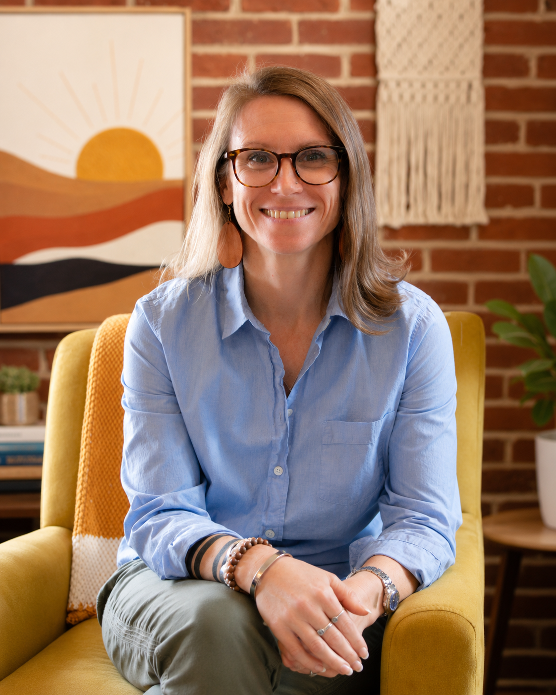 A woman with glasses, long brown hair, and earrings is smiling and seated on a yellow chair in a cozy room with a brick wall and decorative items in the background.