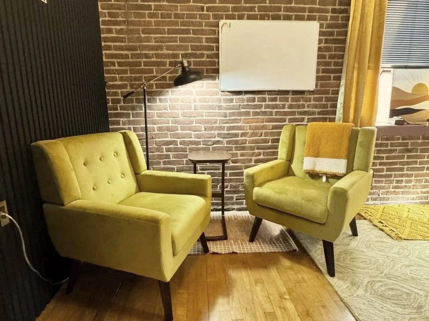 A cozy seating area with two olive green armchairs, a small wooden table between them, a black floor lamp, a whiteboard on a brick wall, and a window with a landscape painting and yellow curtains.