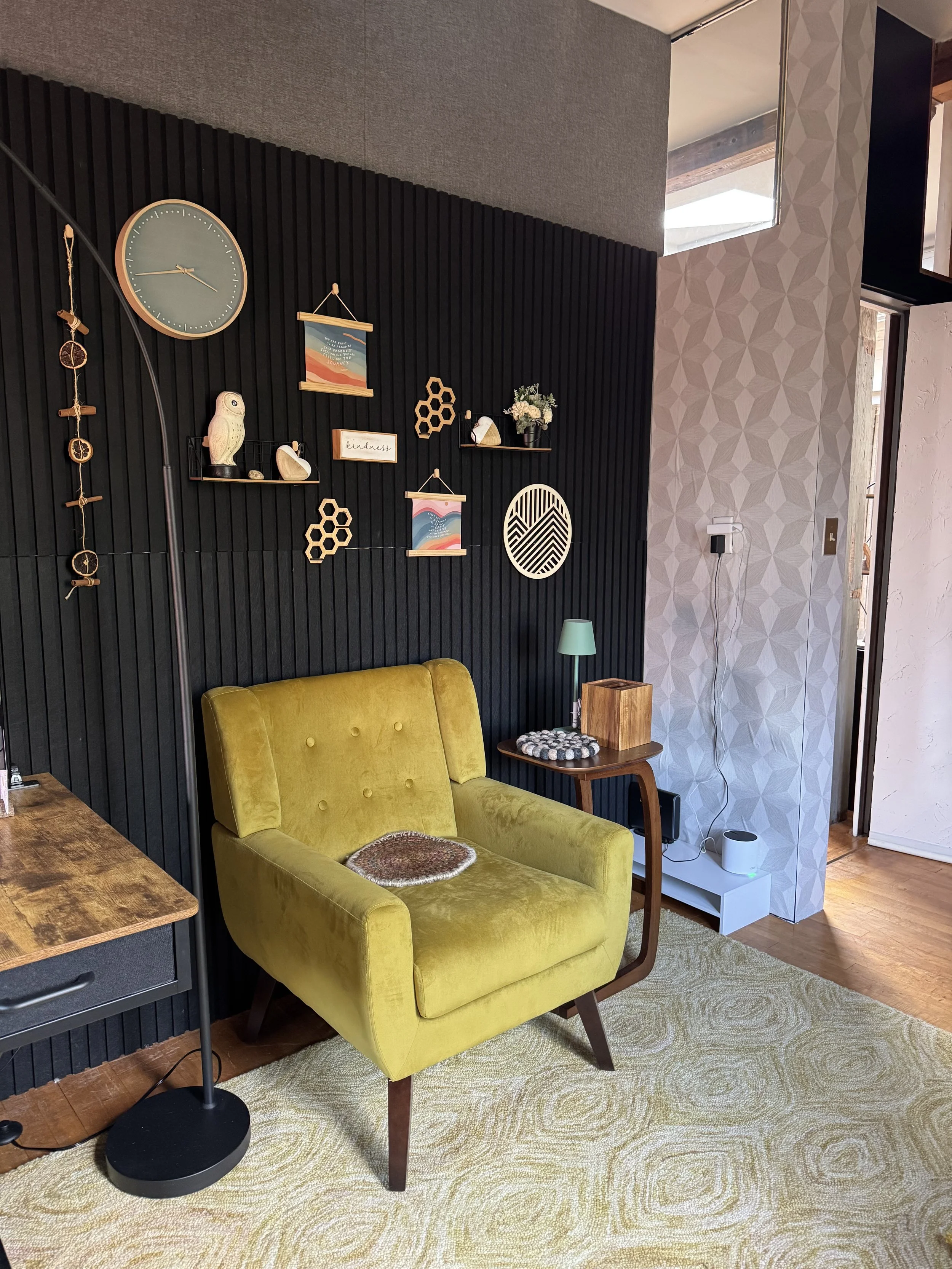 Warm and comfortable psychiatric office with a yellow velvet sofa, a black wall with various decorative items, a clock, framed artwork, and a small side table with a lamp and a wooden box. Light from windows on the right illuminates part of the room.