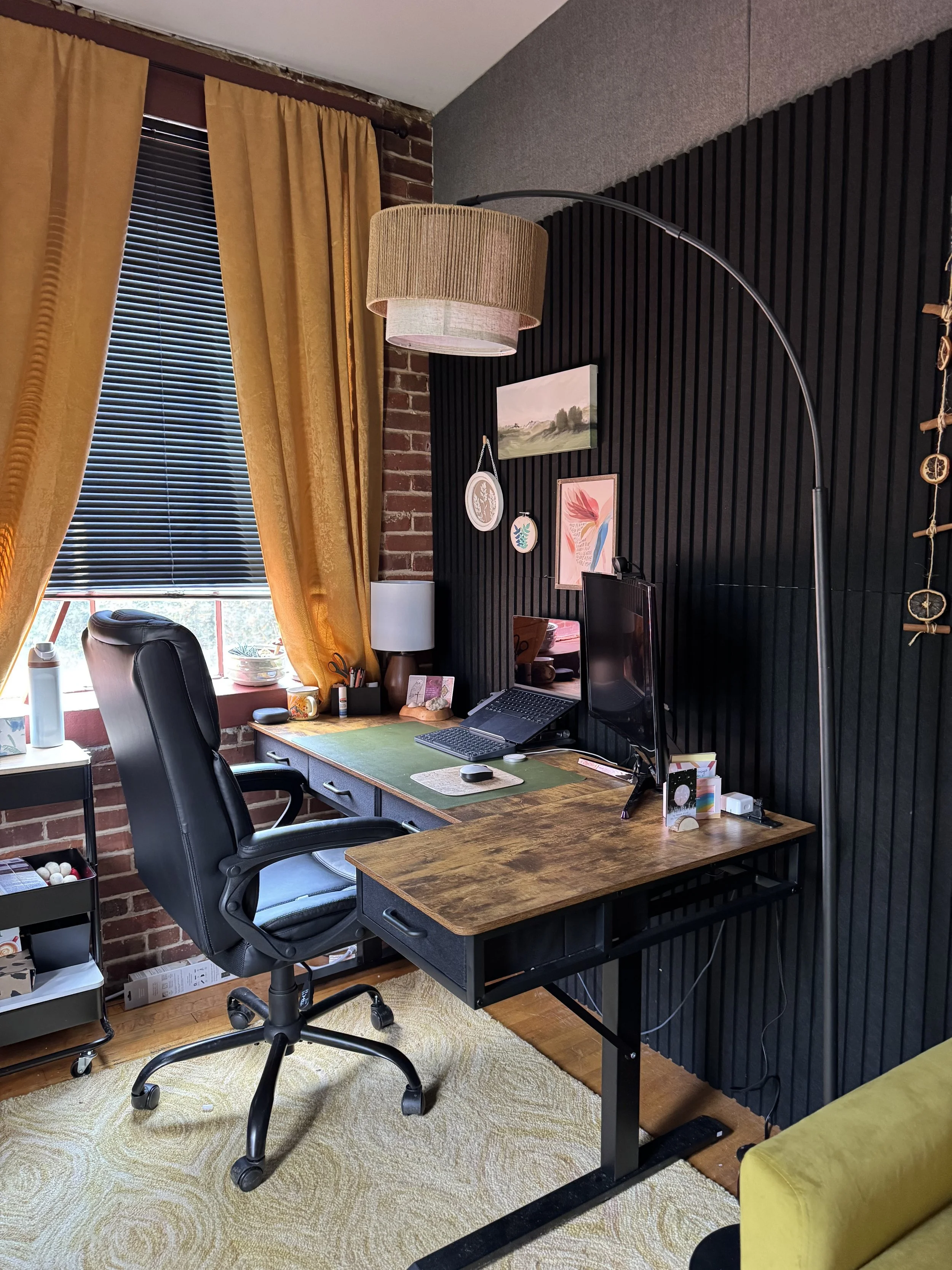 A wooden desk that holds a computer monitor, a laptop, and office supplies. The desk is placed against a black slatted wall with framed artwork and hanging decorations. To the left, there is a window with black blinds and gold curtains, and a small table with a water bottle, cups, and items underneath. The floor is wooden with a light-colored rug.