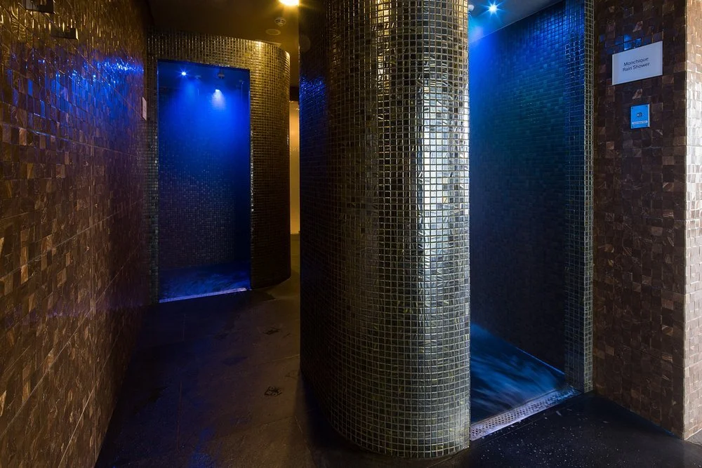 Two shower stalls in a spa with mosaic tile walls and blue lighting.