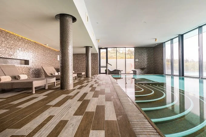 Indoor swimming pool area with lounge chairs, large windows, and a view of water outside.