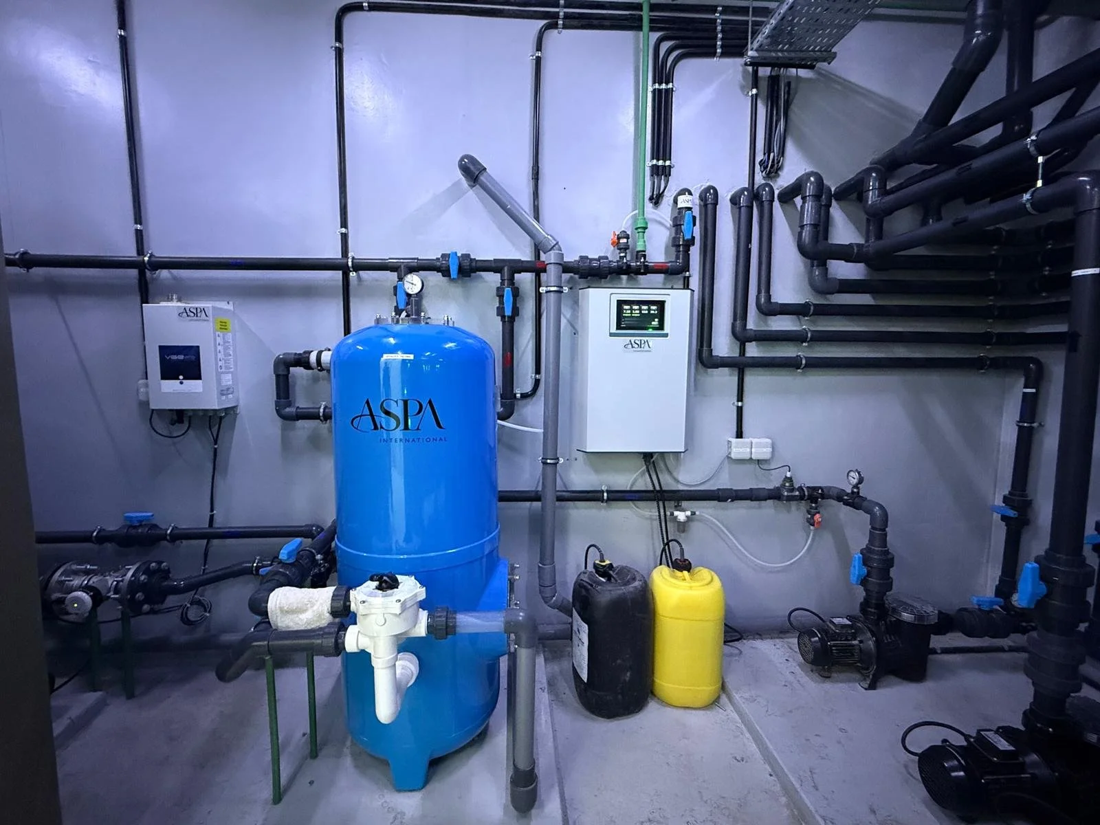 A mechanical room with black piping, a blue water filter tank labeled ASPA, and various equipment including control panels, gauges, and yellow and black tanks.