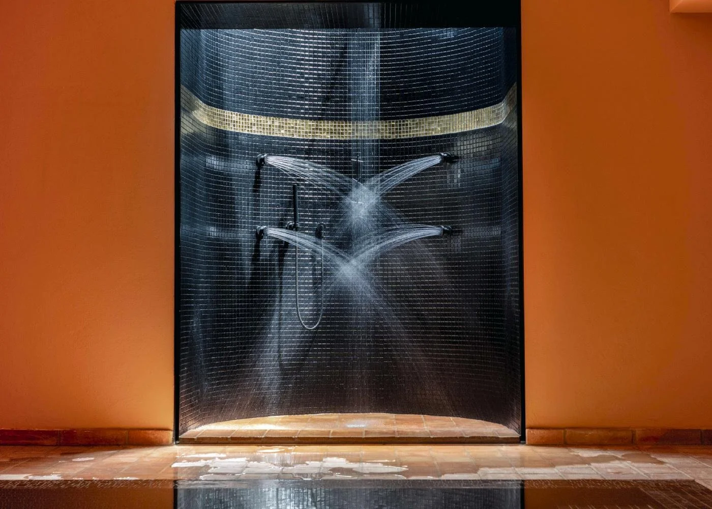 A shower with water spraying from multiple nozzles behind a glass door, situated against an orange wall with a tile floor.