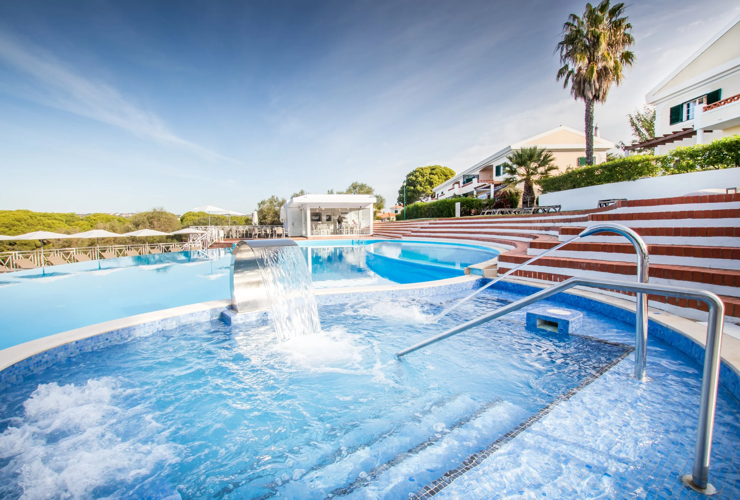 A swimming pool with a waterfall feature, surrounded by white and blue tiled edges, with water cascading into it. In the background, there are white buildings, palm trees, lounge chairs, umbrellas, and a small white cabana. The sky is clear with a few wispy clouds.