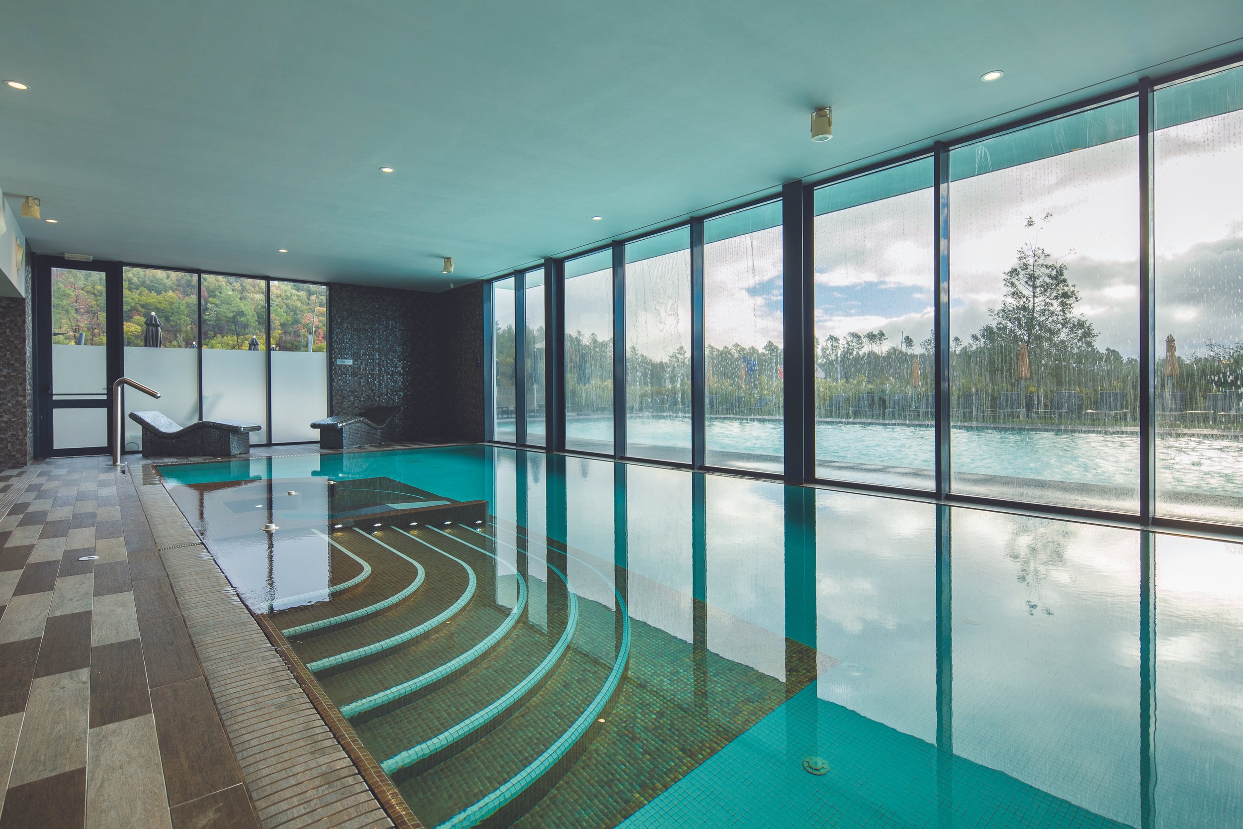 Indoor swimming pool with large floor-to-ceiling windows showing an outdoor landscape with trees and cloudy sky.