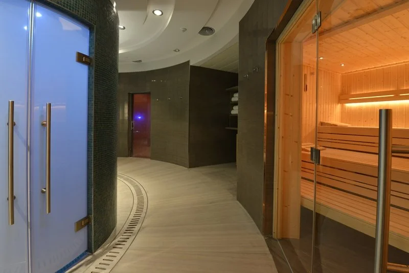 Indoor spa area with a sauna on the right and a curved hallway leading to a door on the left. The sauna room has wooden interior and glass door, while the hallway has dark walls and a light-colored floor.