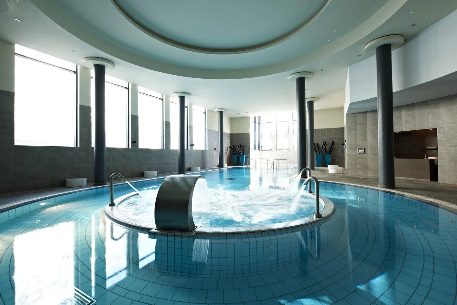 Indoor spa pool area with large windows, water jets, and steps, featuring a modern design with columns and tiled floors.