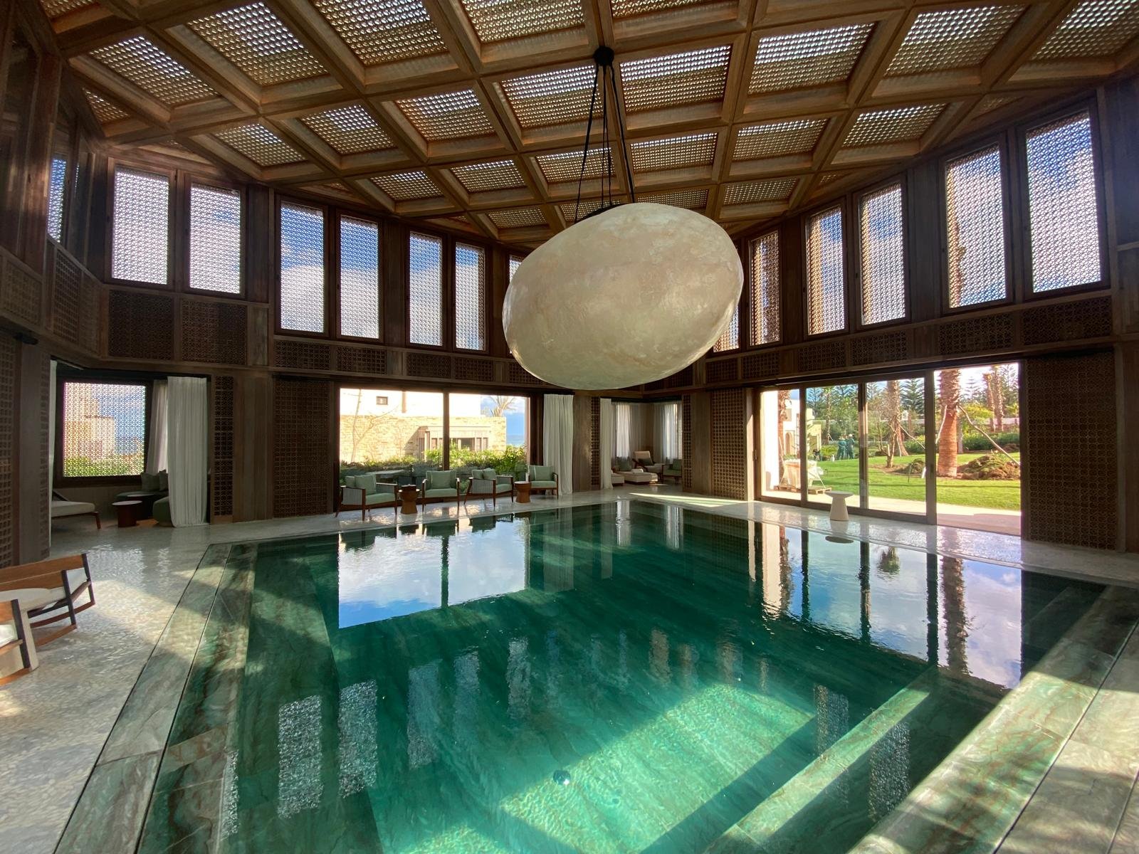 Indoor swimming pool room with wooden lattice ceiling and large windows showing outdoor greenery, giant moon-shaped lamp hanging from the ceiling, and lounge chairs along the walls.