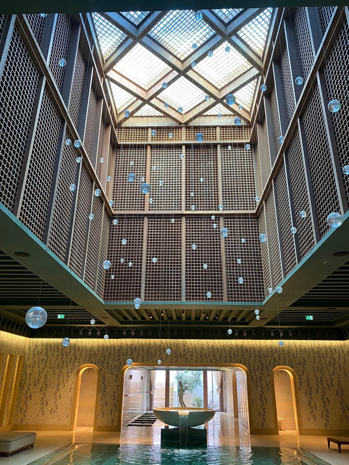 Interior of a building with a tall, open ceiling featuring a skylight, hanging glass orbs, a decorative wall with a honeycomb pattern, and a fountain in the center.