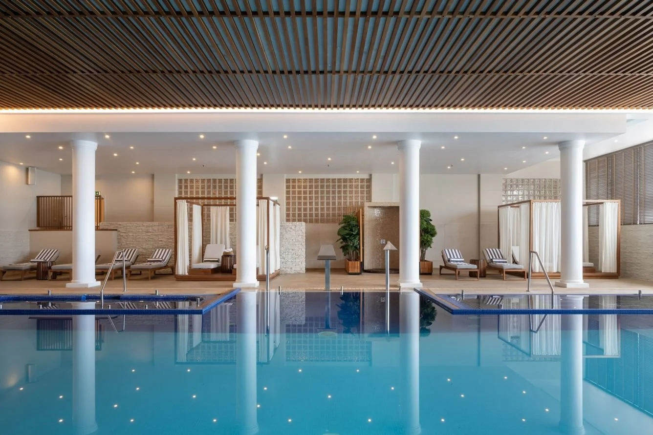 Indoor swimming pool in a modern hotel or spa, with poolside lounge chairs, cabanas with curtains, and decorative plants.