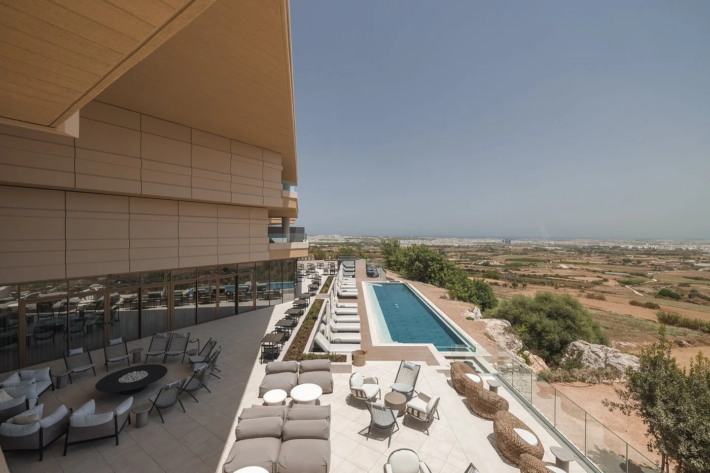 Luxury outdoor pool area with lounge chairs, umbrellas, and seating, overlooking a landscape of fields and distant city skyline.
