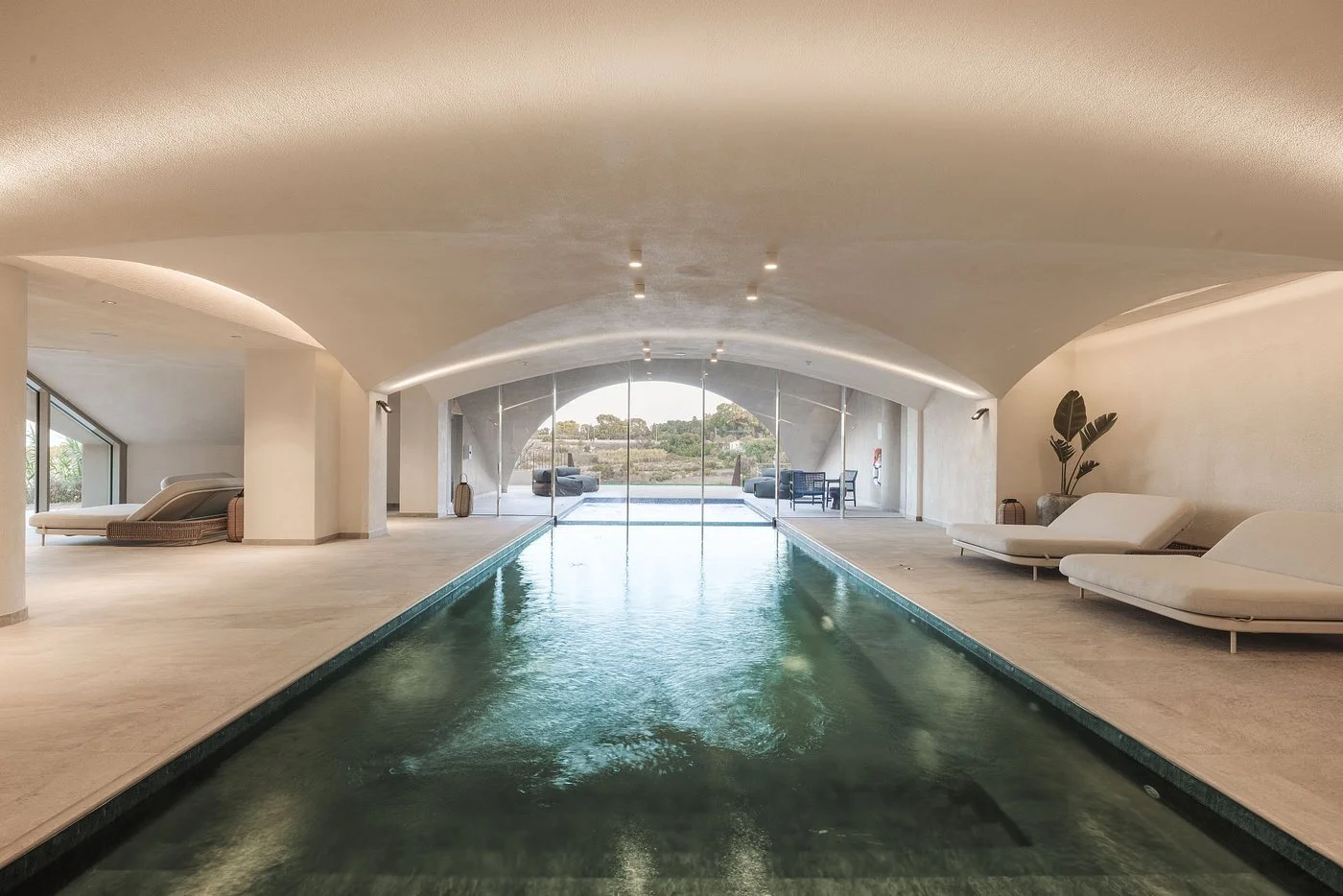 Indoor swimming pool area with white lounge chairs and potted plant, large glass doors leading outside, modern minimalist decor.