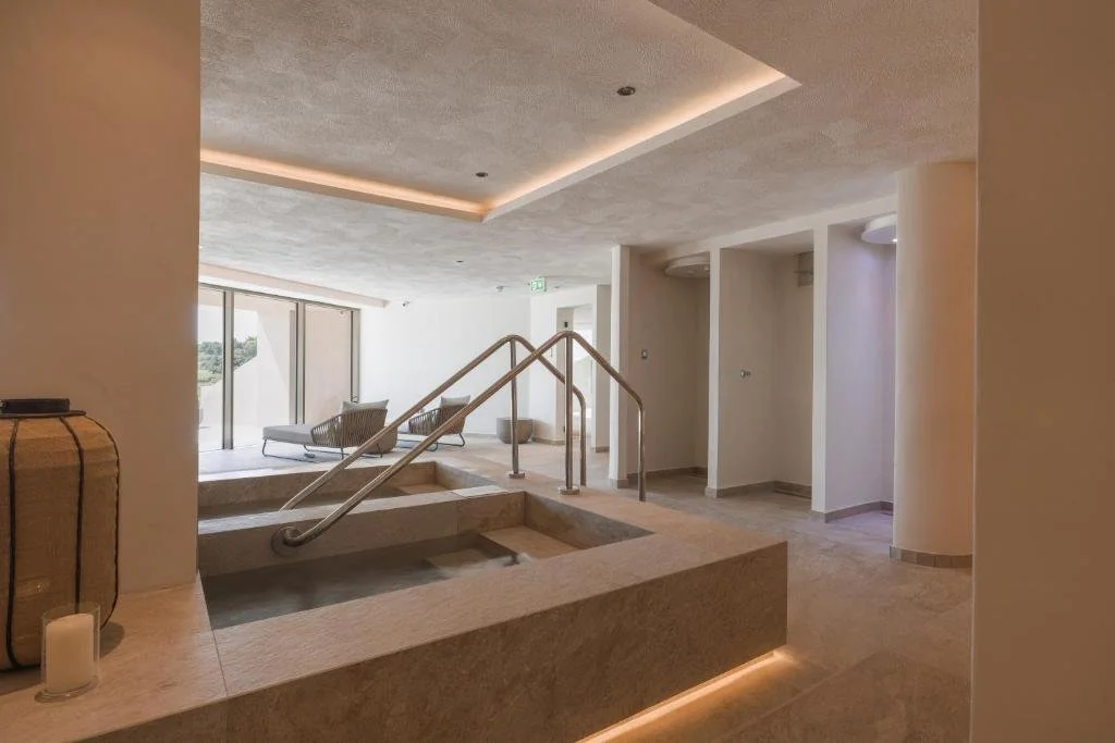 Interior of a modern, minimalist spa or hotel lounge area with a hot tub, lounge chairs, large windows, and soft ambient lighting.