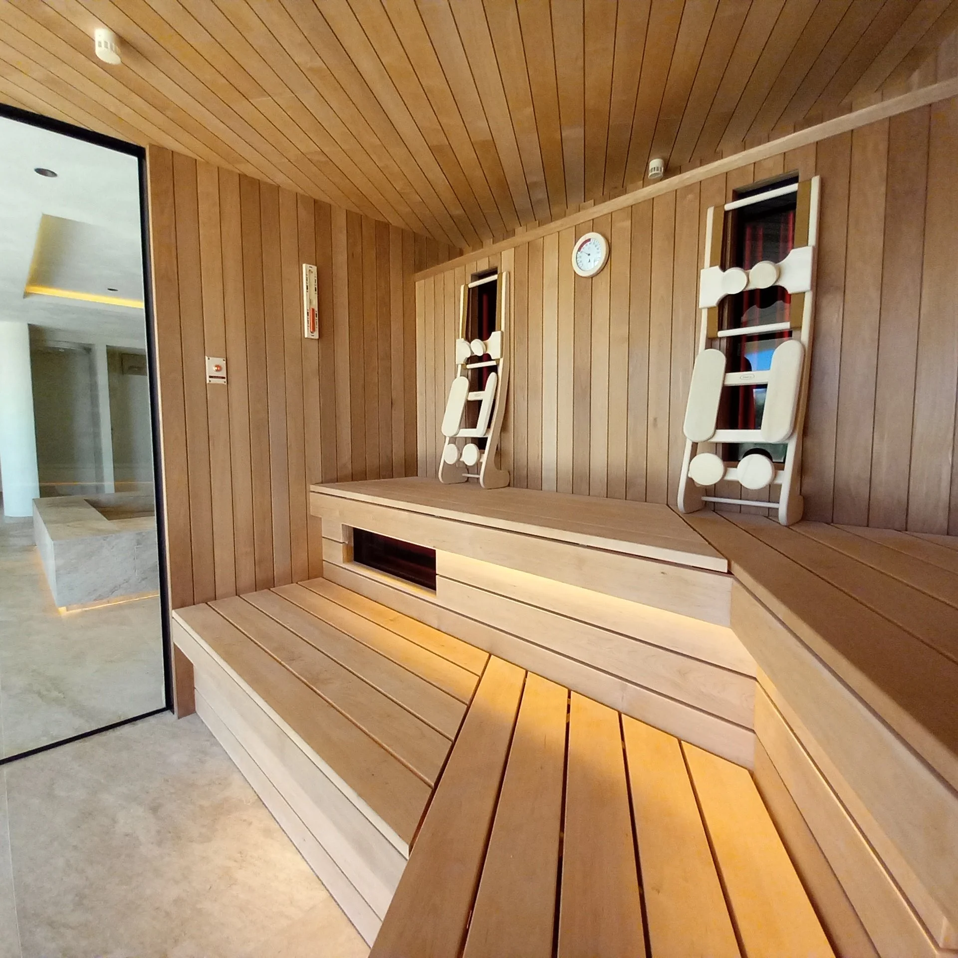 Indoor wooden bench with two white wooden chairs stacked on top of it, wood-paneled walls, a wall clock, and windows with red curtains.
