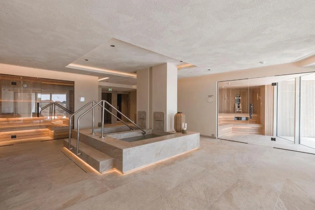 Modern spa interior with a central hot tub, wooden seating area, and glass doors leading outside.