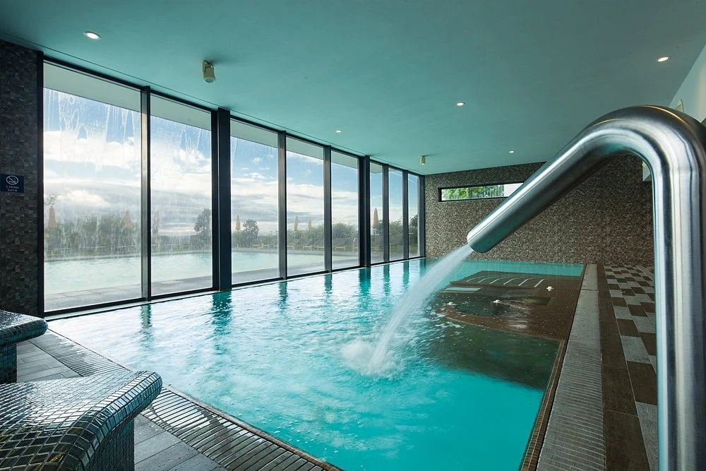 Indoor swimming pool with a large glass window showing an outdoor pool and cloudy sky.