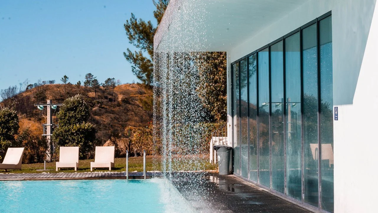 Modern house with large glass windows next to a swimming pool, with a water feature spilling water from a wall, and outdoor chairs in a scenic landscape with hills and trees.