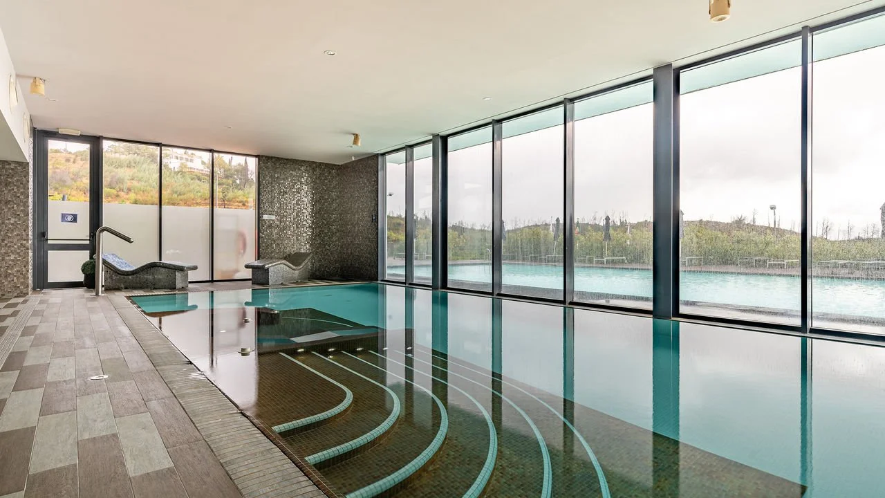 Indoor swimming pool with large glass windows showing an outdoor pool and landscape, featuring pool benches and steps into the pool.
