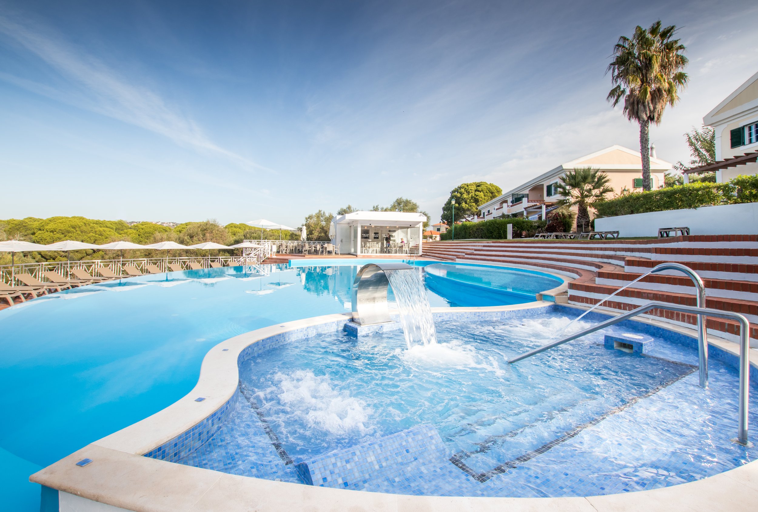 Outdoor swimming pool with blue water, surrounded by lounge chairs, umbrellas, and greenery, under a clear blue sky.