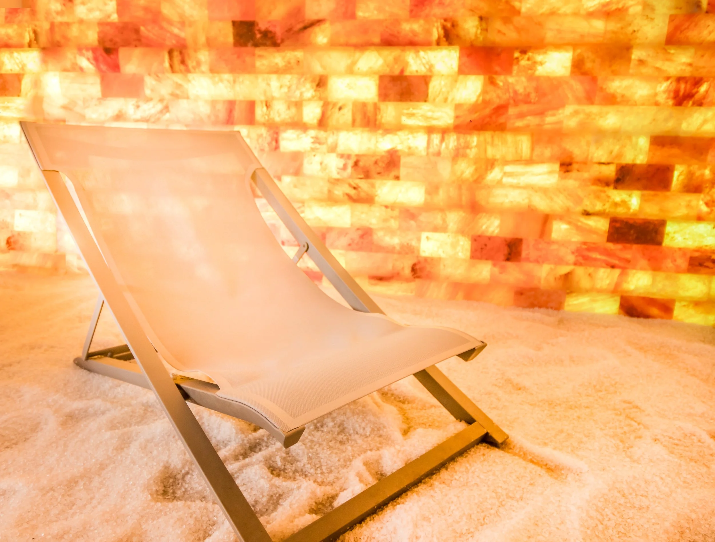A beige lounge chair with a metal frame placed on a salt or salt-like floor against a saltbrick wall that emits a warm orange glow.