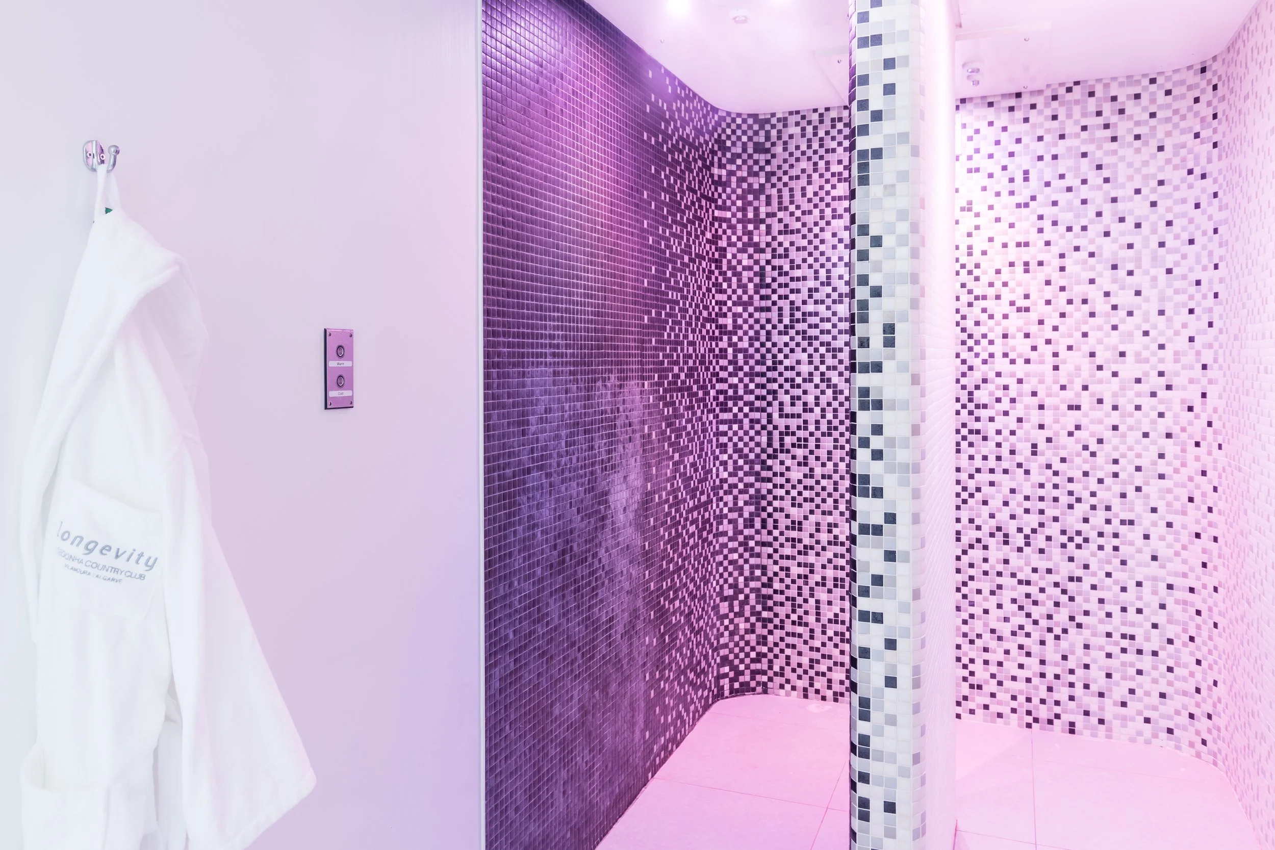 A shower with purple tiles in a gradient pattern from dark to light, with a curved corner and a tiled column with a checkered pattern. A white towel with text hangs on a hook on the left wall, and there's a pink-hued lighting.