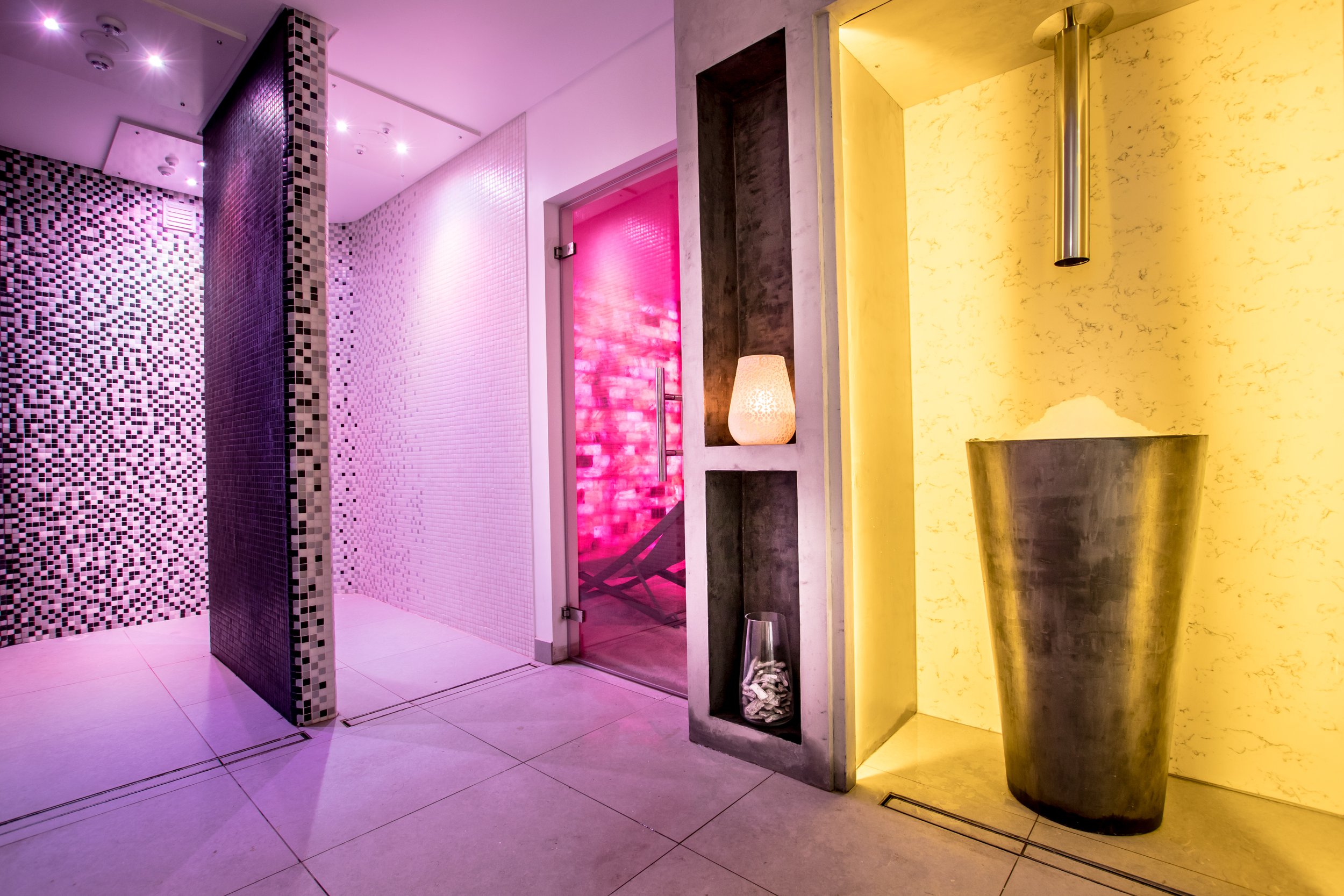 Interior of a modern spa or wellness center with colorful lighting, mosaic tiled walls, a glass door, and decorative elements like a lamp and a large vase.