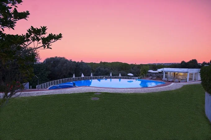 An outdoor swimming pool at sunset, surrounded by a grassy lawn and trees, with a modern pool house in the background.