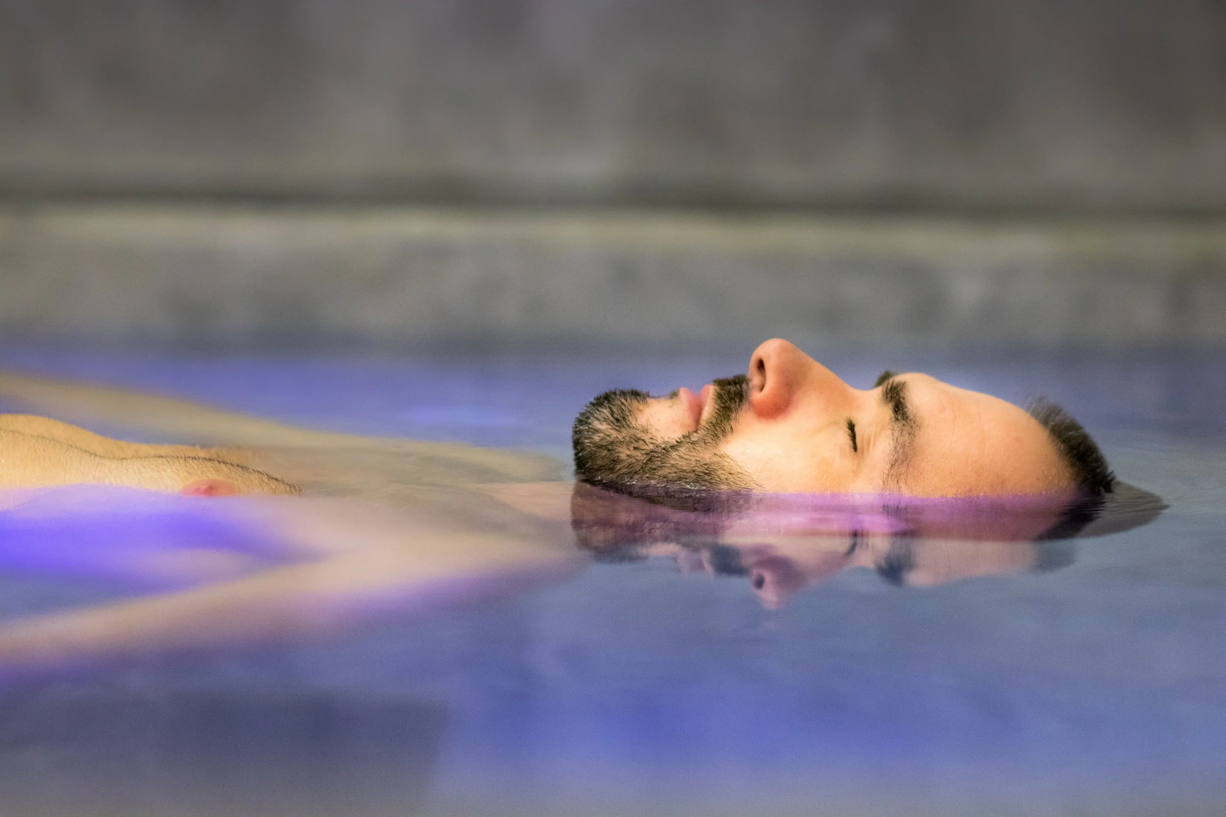 A man with a beard is floating on his back in a body of water with his eyes closed.