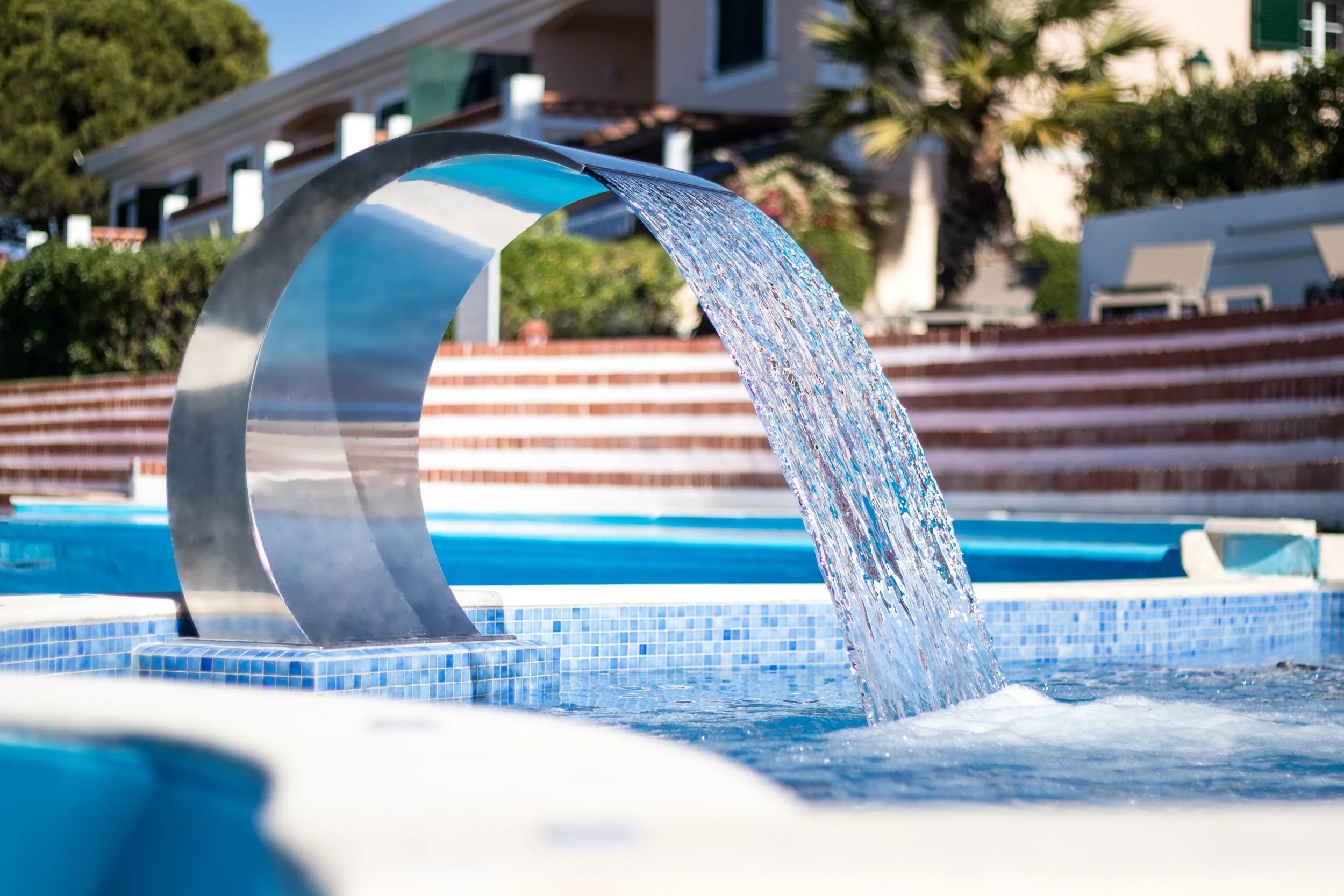 Close-up of a modern fountain with water flowing in a pool with blue tiles, in an outdoor setting with residential buildings, trees, and a sunny sky in the background.