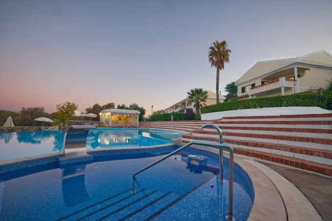 Swimming pool with steps and a metal handrail, surrounded by brick stairs, with a white building and palm trees in the background during sunset.