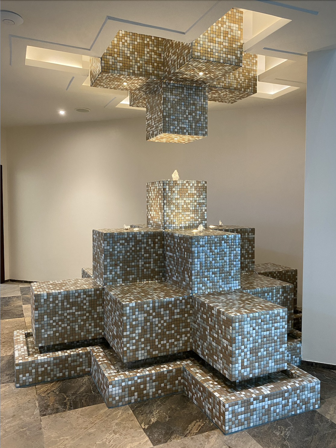 A decorative indoor fountain with mosaic tiled blocks forming a tiered structure, topped with small rocks, and a modern ceiling with built-in lighting.