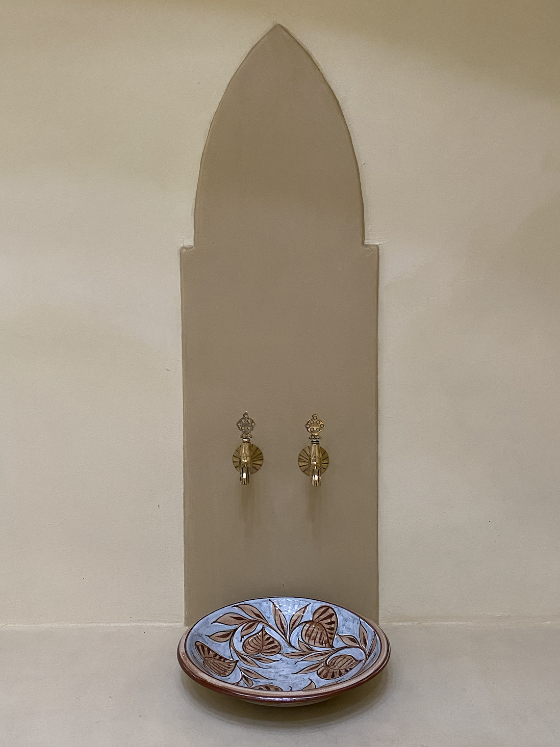 Decorative wall with two gold hooks, a mirrored arched shape, and a ceramic dish with leaf patterns at the bottom.
