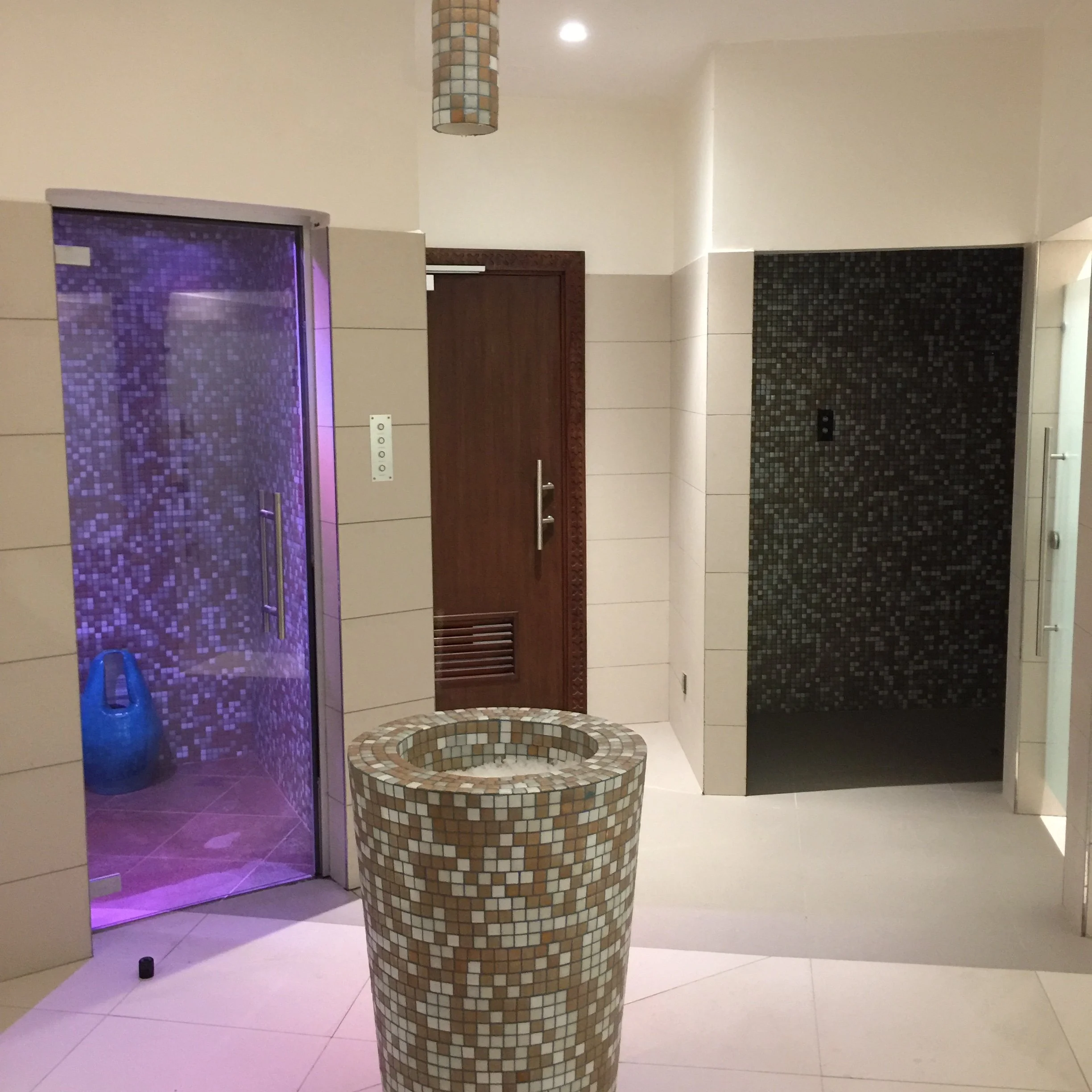 A mosaic tiled fountain with a circular basin in a modern interior space, with a purple-lit shower area to the left and a wooden door behind the fountain.