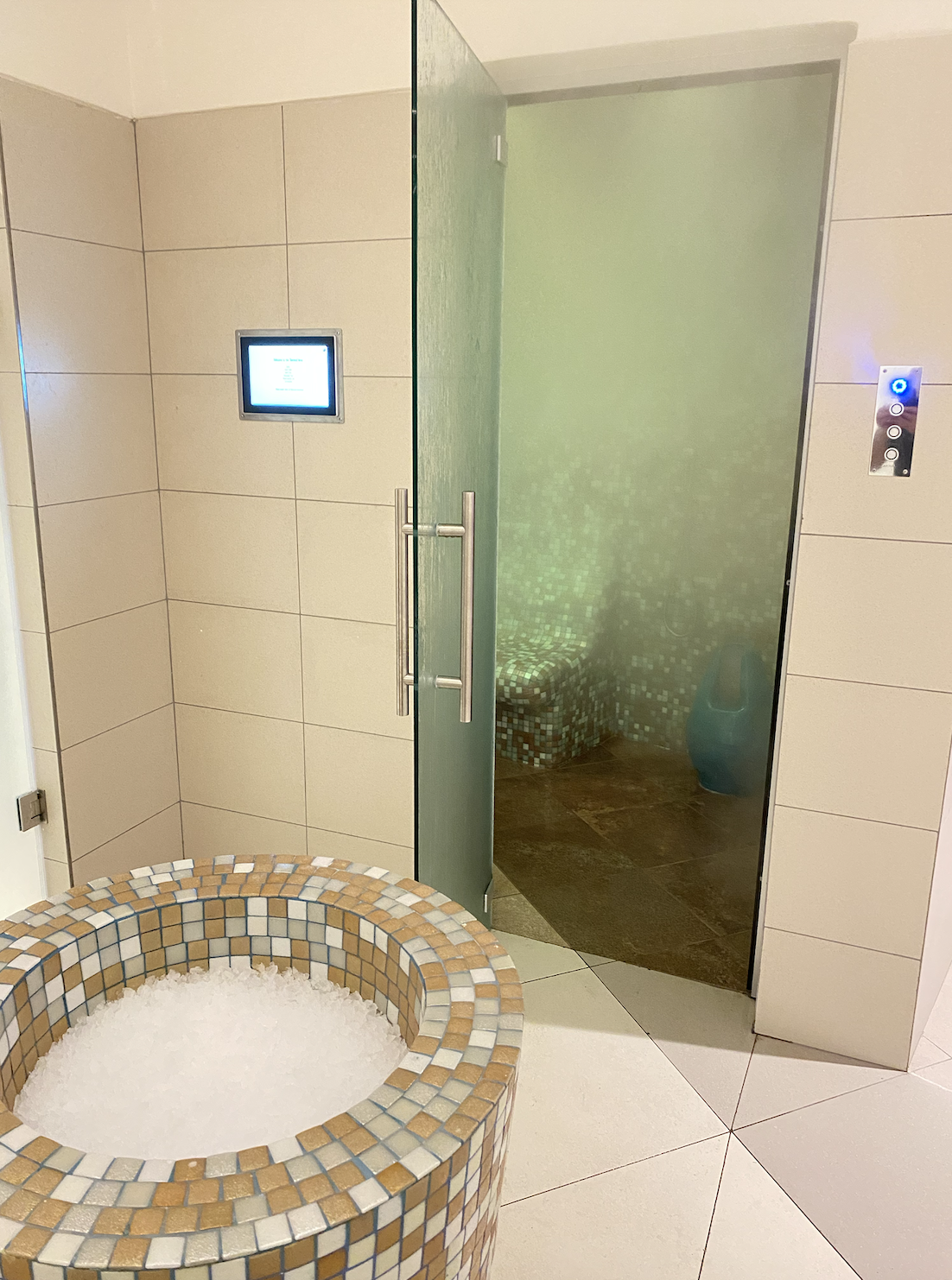 A tiled room with a bubble-filled whirlpool tub in the foreground and a glass door leading to a steam room or sauna in the background. The steam room has mosaic tile walls and a bench.
