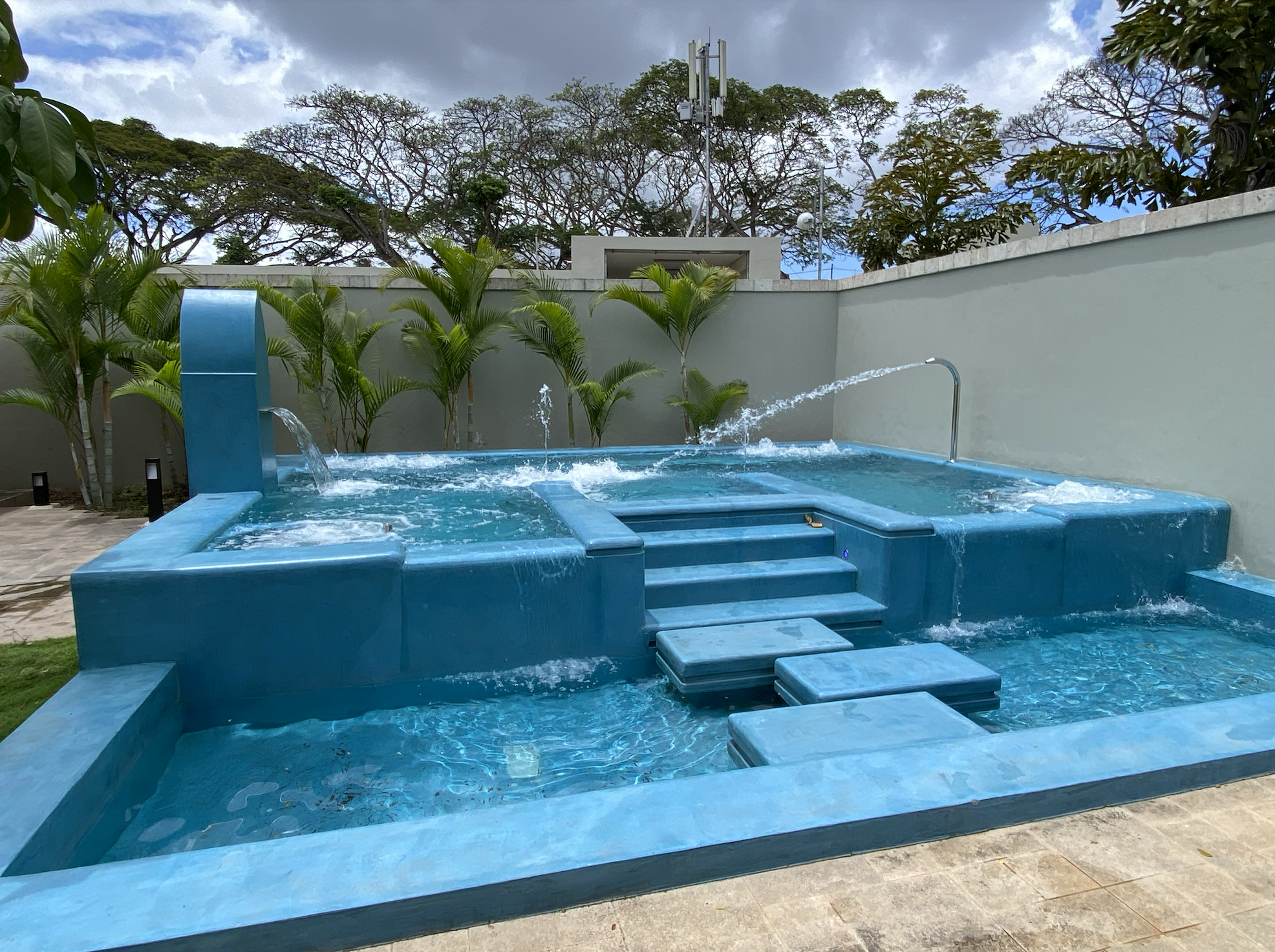A turquoise hot tub with built-in seating and a waterfall feature, surrounded by lush green plants and a gray wall, with trees and cloudy sky in the background.
