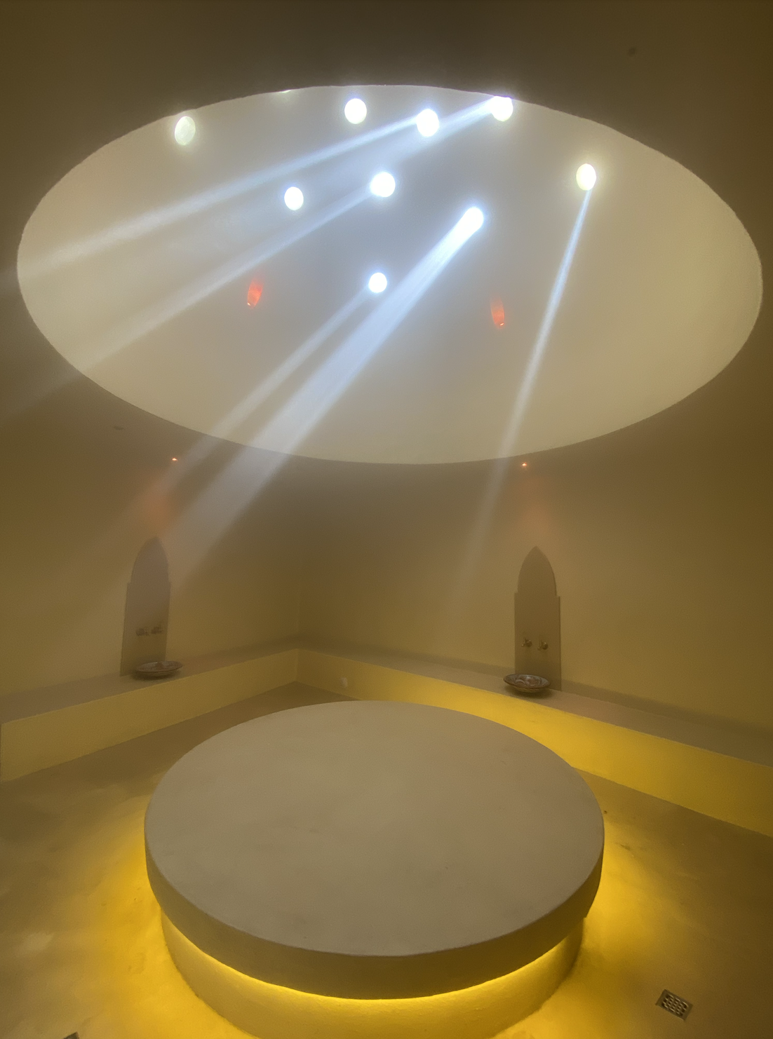 A room with a round bedding platform illuminated by yellow lighting, a circular skylight overhead with multiple white and red spotlights creating beams, and small alcoves on the walls with decorative elements.