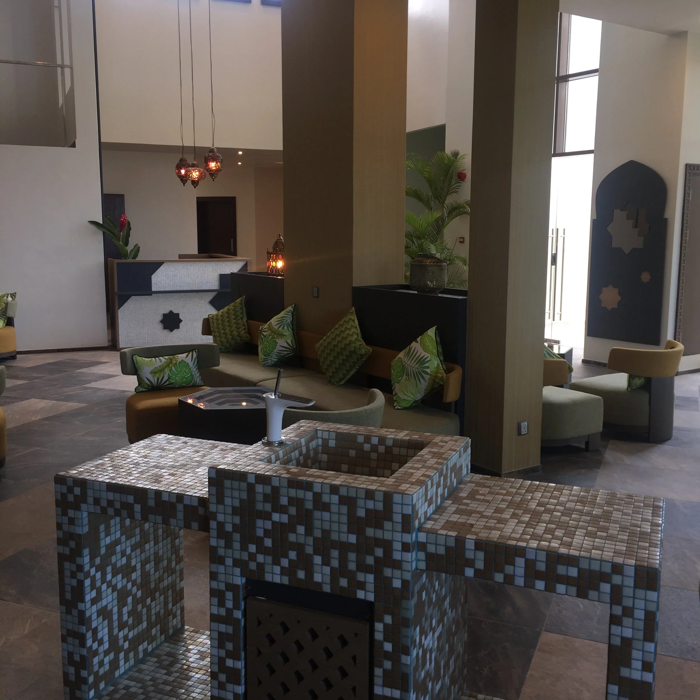 Indoor space with modern decor featuring a mosaic-tiled table in the foreground, cushioned seating with patterned pillows, a plant, decorative lanterns, and large windows allowing natural light.