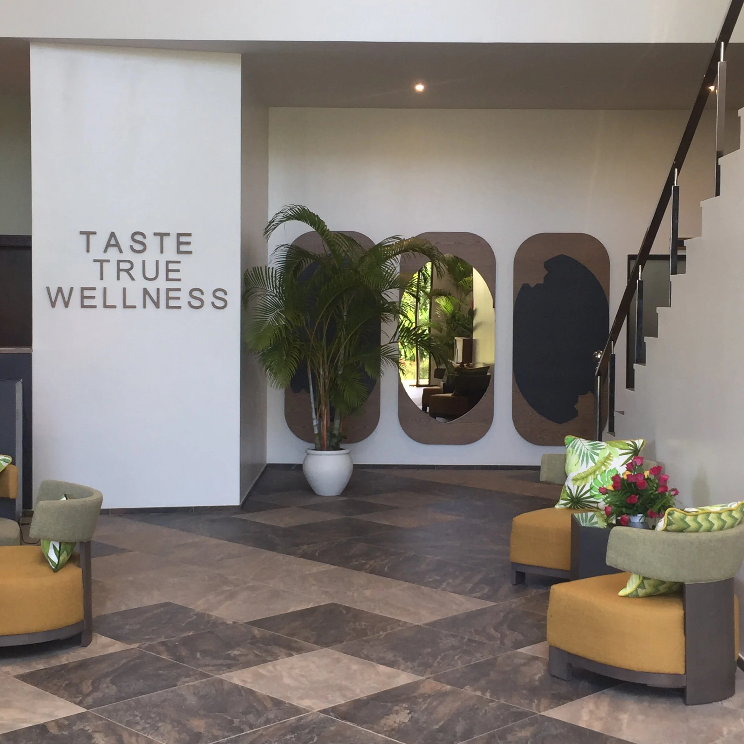 Interior of wellness center with white walls, a display with the words 'Taste True Wellness,' a large white vase with green plants, round seating with patterned cushions, and a staircase with black railing.