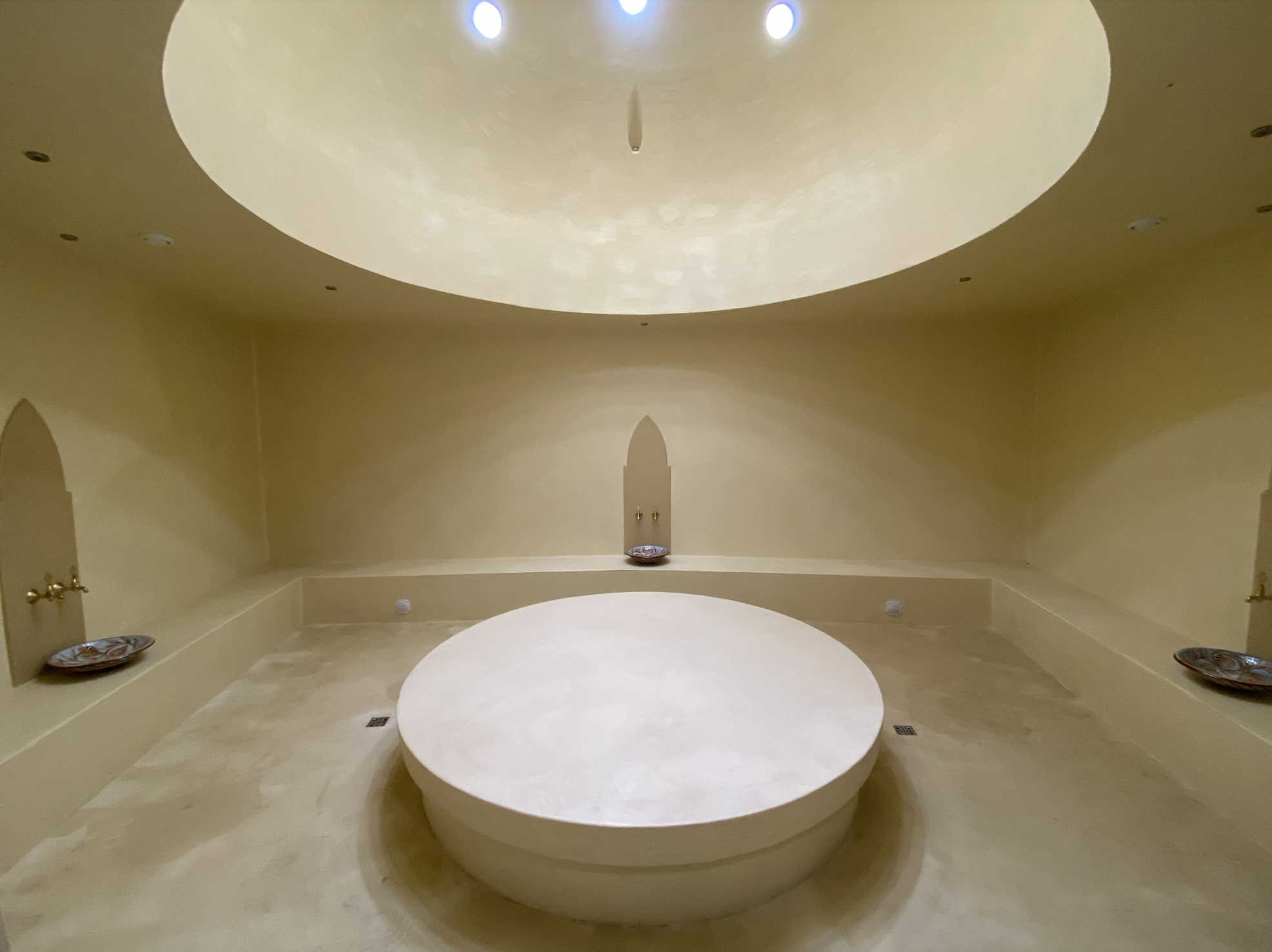 A luxurious spa room with a large, circular sunken bathtub at the center, surrounded by beige tile flooring and pale yellow walls, with decorative plates on the sides and small lights on the ceiling.