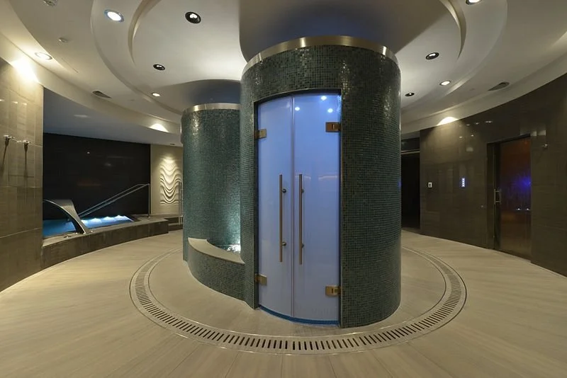 A modern indoor spa with a circular glass shower enclosure, tiled walls, and a sauna area with sitting benches and water feature.