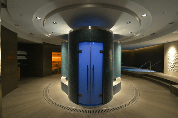 Modern spa or wellness center with a central circular blue door, surrounded by dark textured walls, with a relaxed seating area to the right, and ambient lighting.