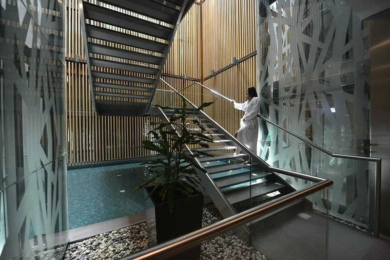 Person in a white robe holding a spray wand, standing on a staircase inside a modern building with decorative glass walls, a water feature, and wooden accents.