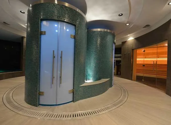 Modern interior space with two cylindrical teal-colored structures, one with glass doors, and a curved seating area with soft lighting, wood-paneled wall in the background.