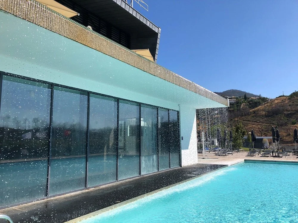 A swimming pool with water cascading down from the building's overhang, reflecting sunlight. The building has large glass panels and is located near a hillside with dry vegetation, under a clear blue sky.