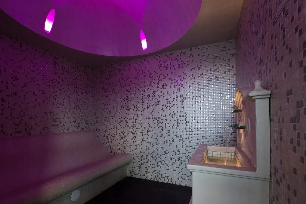 A small, tubular room with mosaic-tiled walls illuminated by purple lights, featuring a pink cushioned bench and a pink sink with a small mirror.