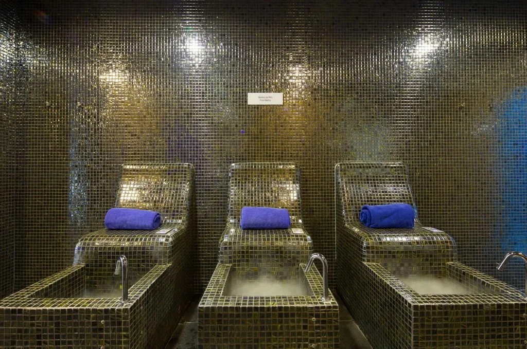 Three metallic massage chairs with blue towels draped over the headrests, placed in front of sinks with steaming water at a spa or bathhouse, with small mosaic tiles on the walls.