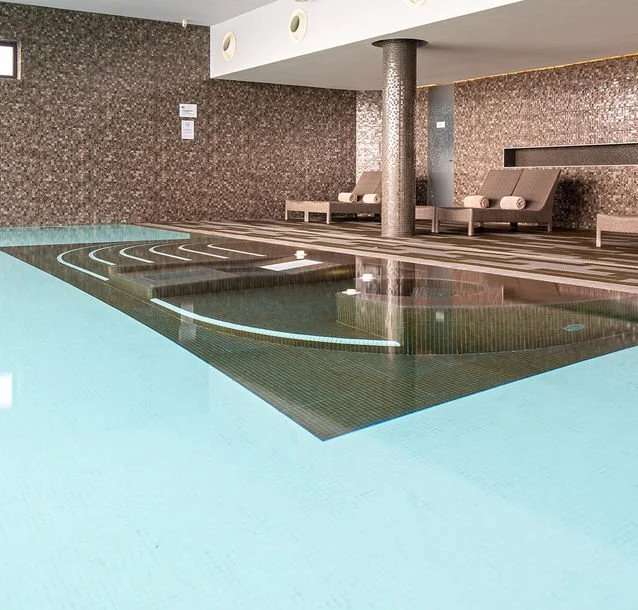 Indoor swimming pool area with steps leading into the pool, lounge chairs along the wall, and a column supporting the ceiling.