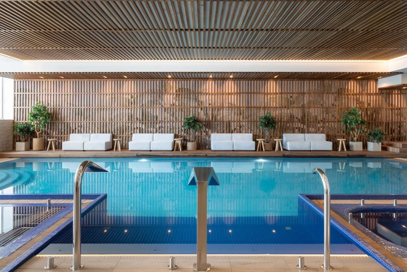 Indoor swimming pool area with white lounge chairs and potted plants along a wooden wall, with a wooden slatted ceiling.